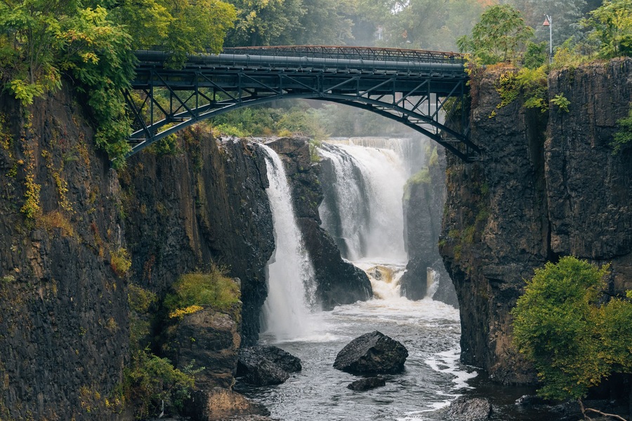 A bridge over a waterfall with surrounding rocky cliffs and greenery.