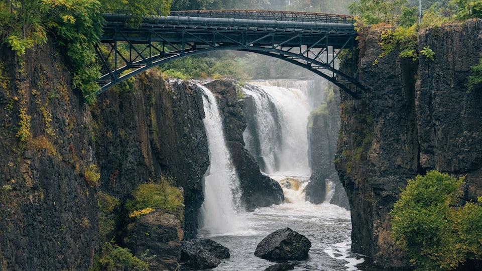 A bridge over a waterfall with surrounding rocky cliffs and greenery.