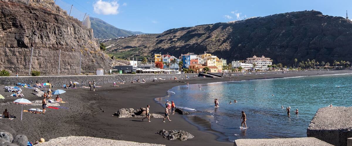 A beach with people sunbathing and swimming, a rocky shoreline, and colorful buildings in the background.