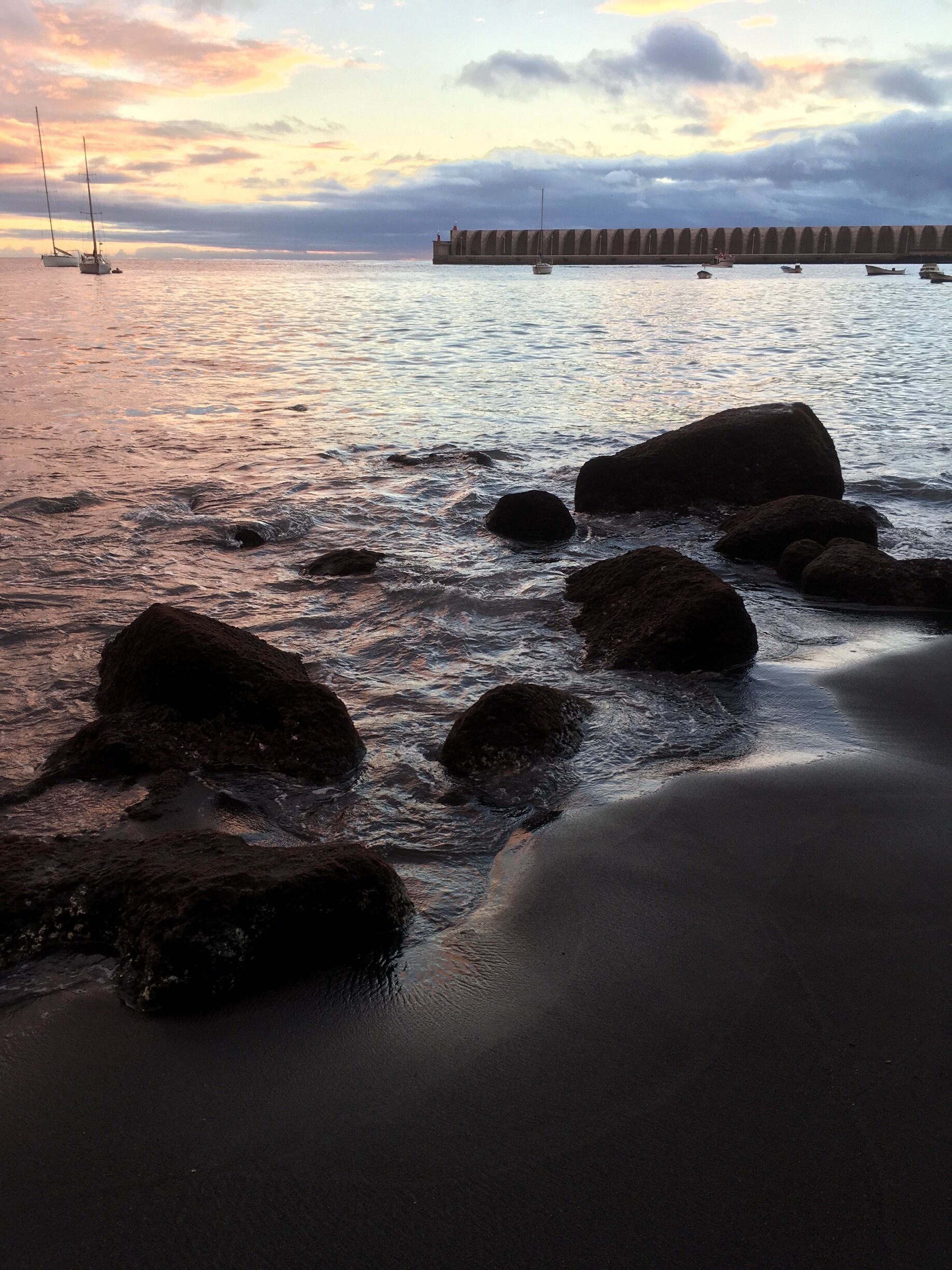 A beach with rocks and calm water, a pier in the background, and a sunset sky.