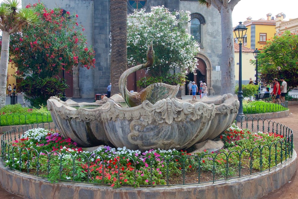 Una gran fuente decorativa con elaboradas esculturas, rodeada de coloridos macizos de flores y un borde de piedra.