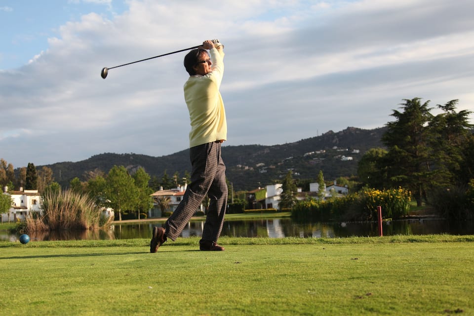 En person spiller golf på en solskinsdag med en sø og bakker i baggrunden.