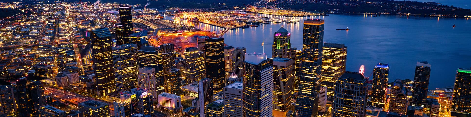A cityscape at dusk with illuminated skyscrapers, a waterfront, and a bridge.