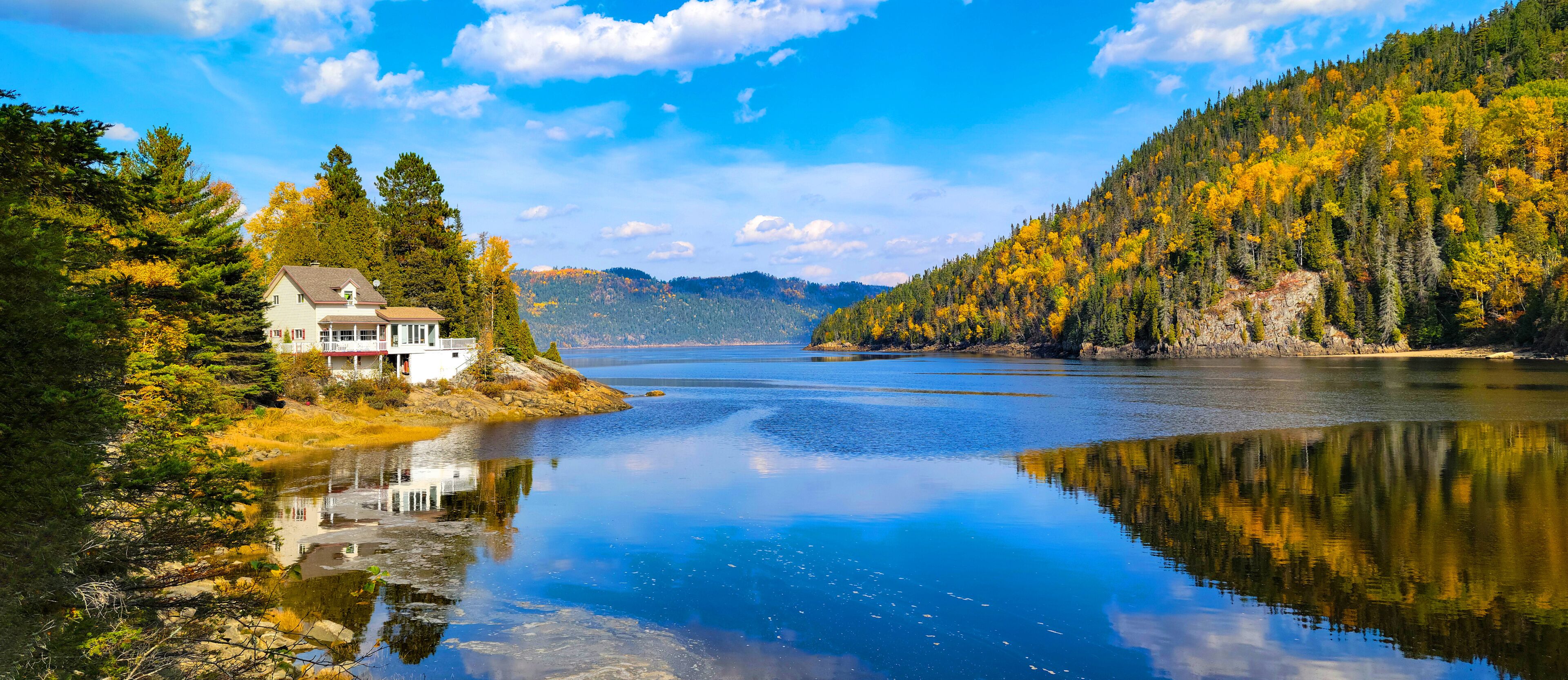 A serene lake with a house on a rocky shore, surrounded by autumn-colored trees and a clear blue sky.
