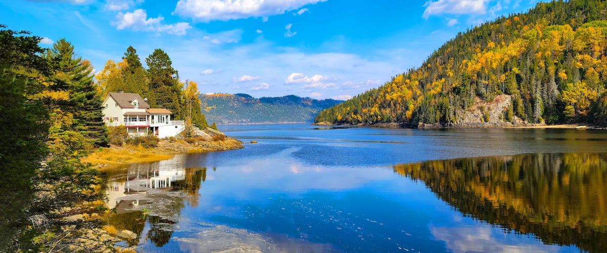A serene lake with a house on a rocky shore, surrounded by autumn-colored trees and a clear blue sky.