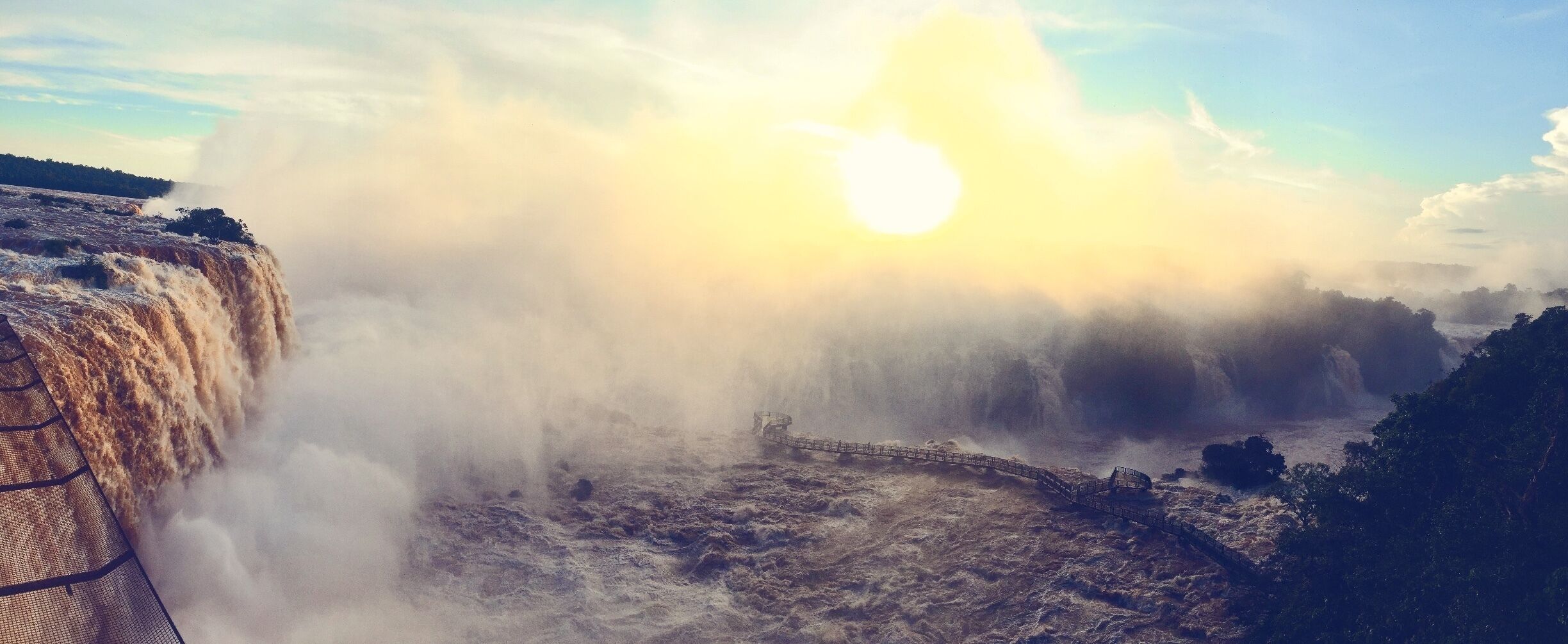 A waterfall with mist rising from the water, a bridge crossing over the turbulent flow, and a sun setting or rising in the background.