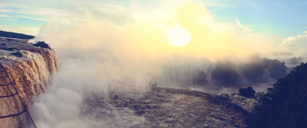 A waterfall with mist rising from the water, a bridge crossing over the turbulent flow, and a sun setting or rising in the background.