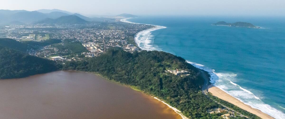 An aerial view of a coastal city with a beach, forested hills, and a large body of water.
