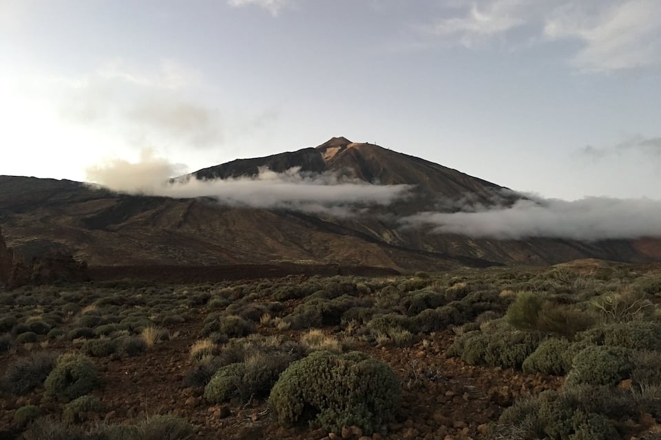 Una montaña con nubes en la cima, rodeada de vegetación escasa.