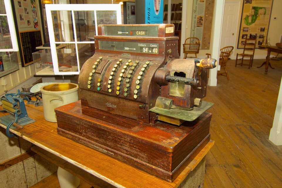 An antique cash register with a wooden base and a brass top, placed on a wooden table in a room with wooden floors and chairs.