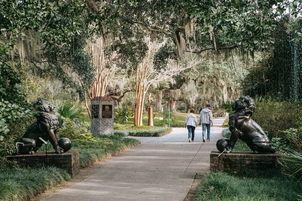 A pathway flanked by bronze statues of lions, leading through a lush garden with trees and hanging moss.