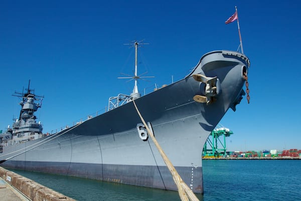 Ein großes Kriegsschiff liegt in einem Hafen, der von einem wolkenlosen, blauen Himmel überdacht wird.