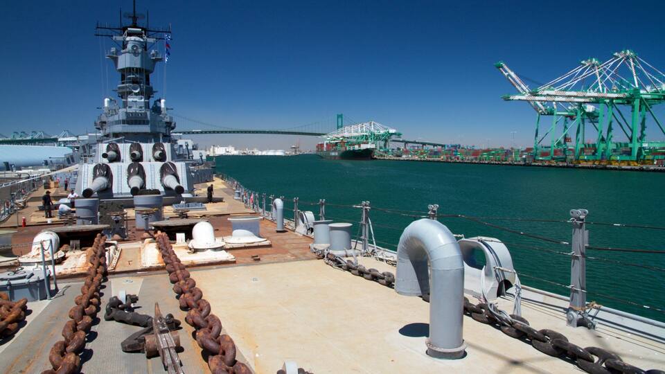 A naval ship docked at a port with a large cargo ship and a green crane in the background.
