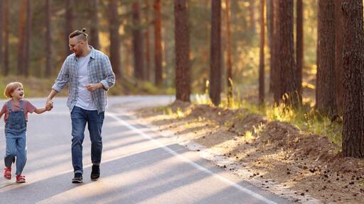 Dad and his little daughter are walking along a forest road among tall pines. Family walk in the forest at sunset, man and little girl. Banner for website header design with copy space.