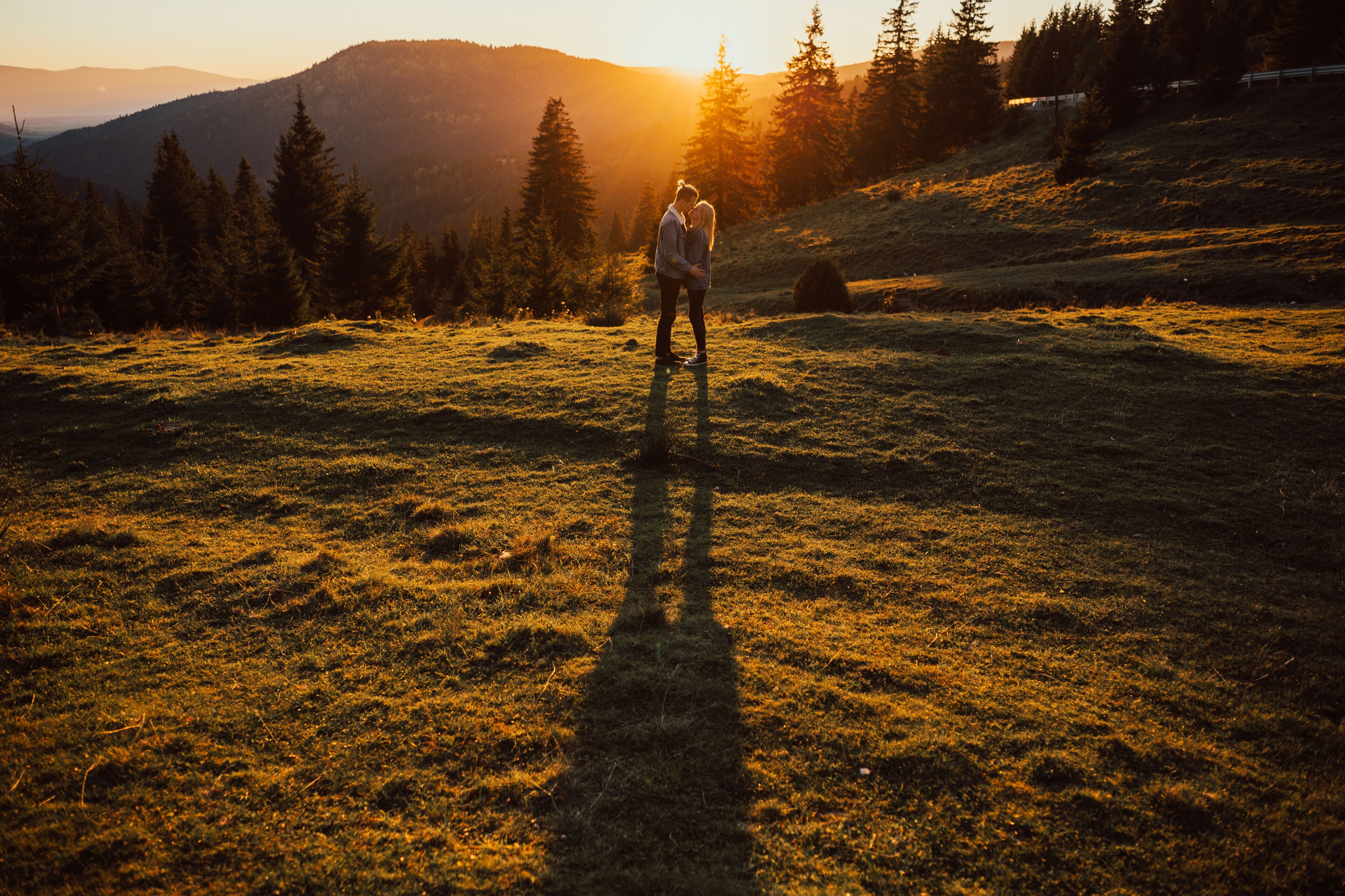 Warm evening sun light. Happy young couple hugging and kissing at sunset with amazing mountain view and pine forest. Silhouette.
