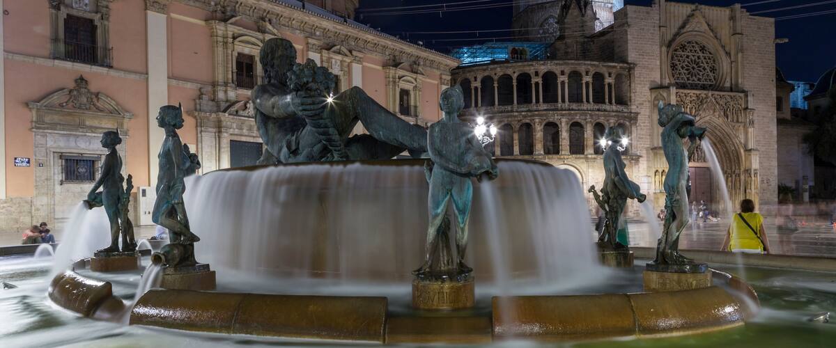 A nighttime view of a historic fountain with statues, surrounded by classical architecture.