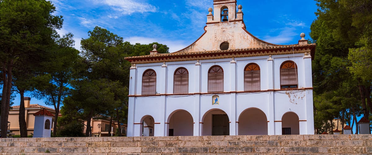 A white church with a bell tower and arched windows, surrounded by trees and stone steps.