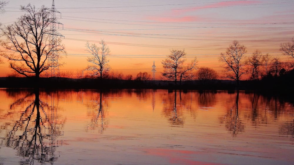 Un coucher de soleil sur un lac paisible, avec les reflets des arbres et des lignes électriques.