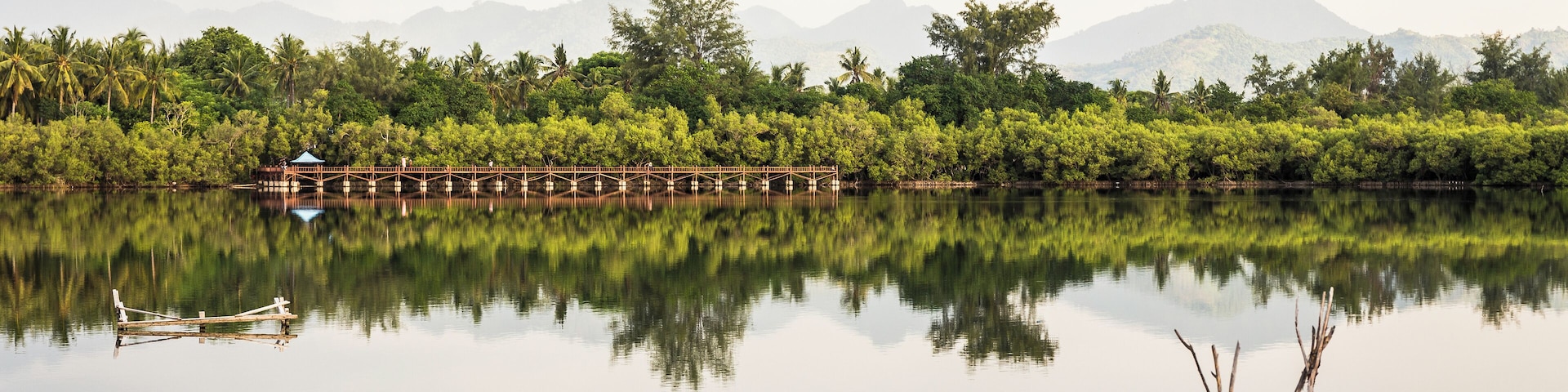 A calm lake with a wooden bridge, a small hut, and a reflection of trees and mountains in the water.