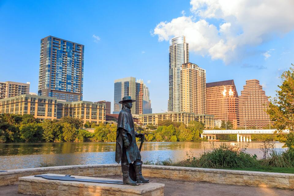 A statue by a river with a city skyline in the background.