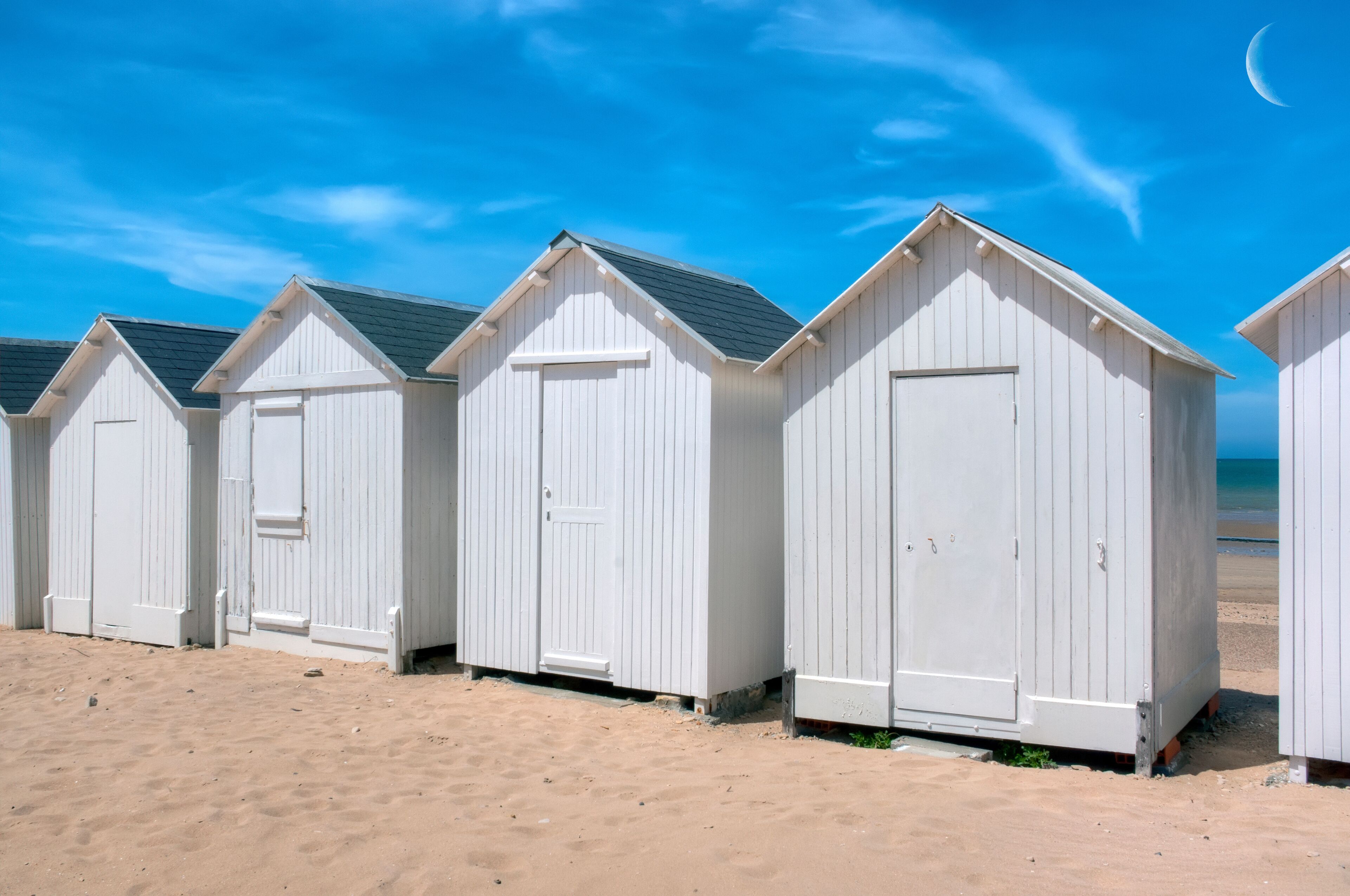 A row of white beach huts on a sandy beach with a clear blue sky and a crescent moon.