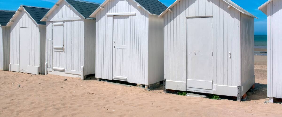 A row of white beach huts on a sandy beach with a clear blue sky and a crescent moon.