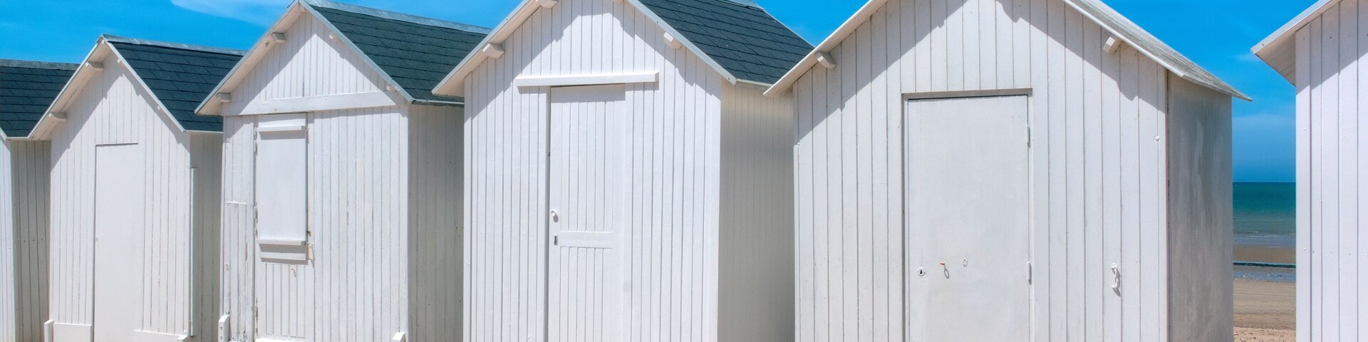 A row of white beach huts on a sandy beach with a clear blue sky and a crescent moon.
