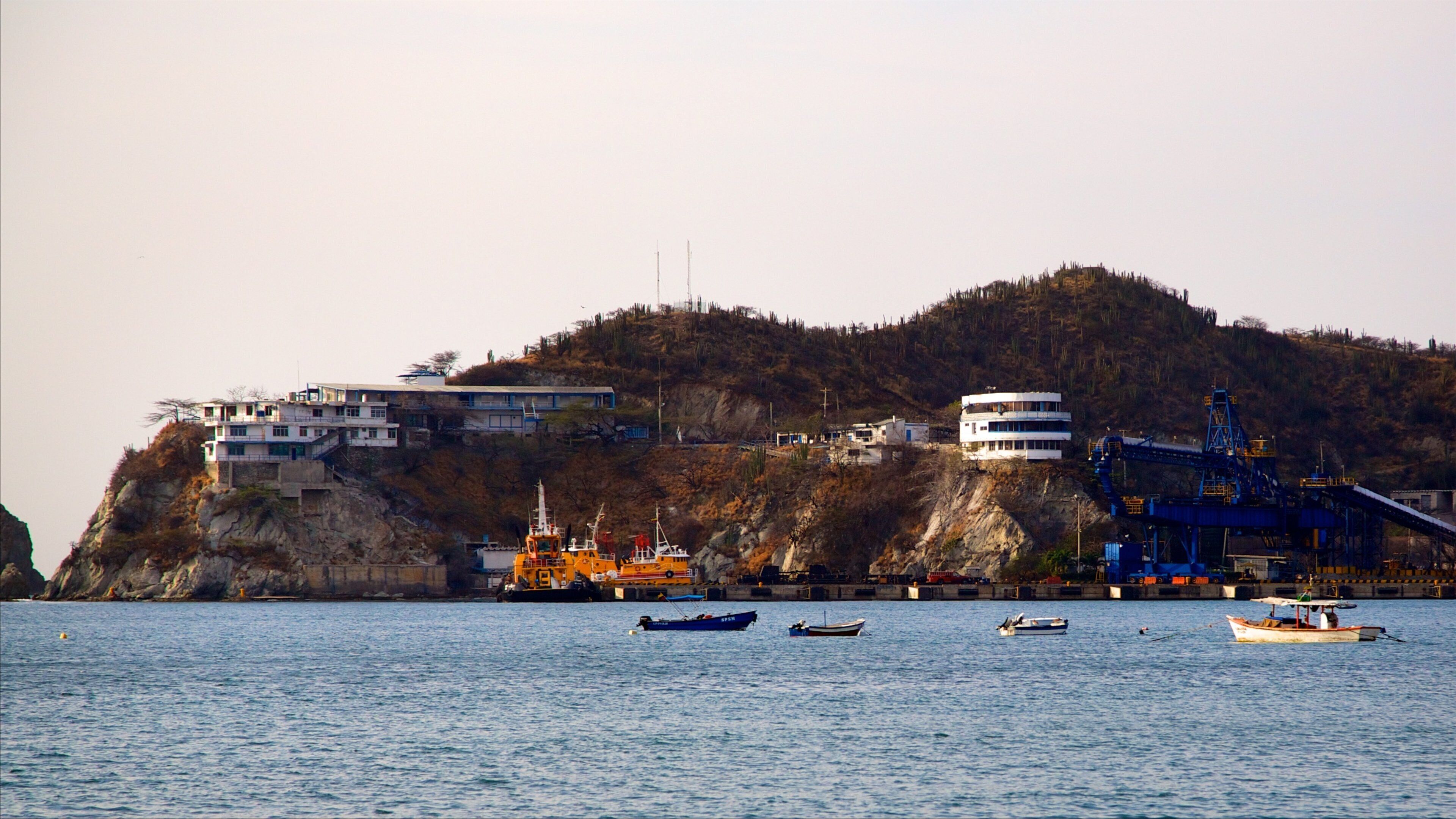 A coastal town with buildings on a cliff, boats on the water, and industrial structures.