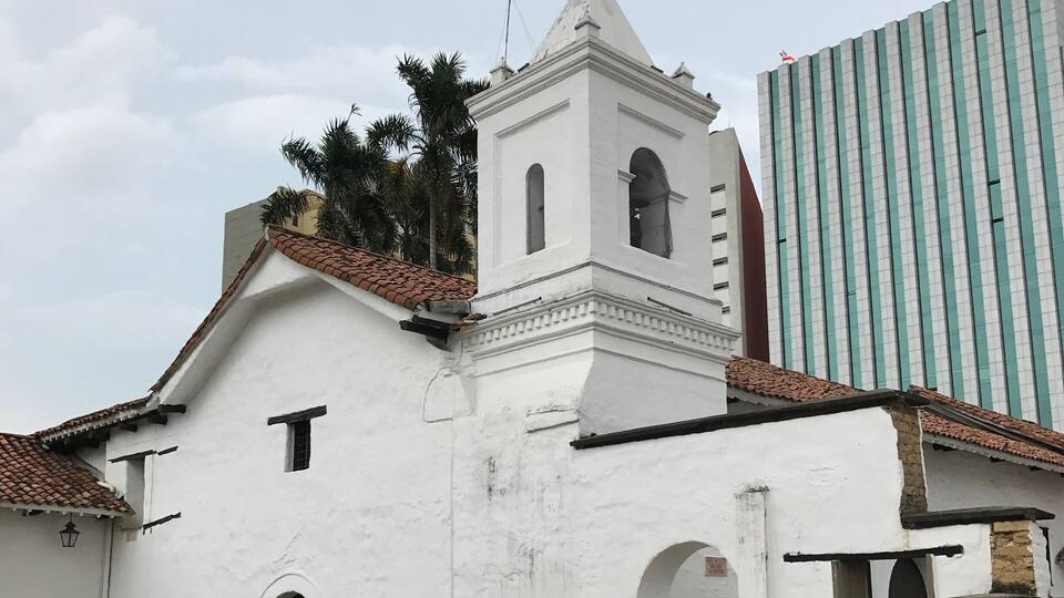A white church with a bell tower and arched doorways, surrounded by cobblestone streets and palm trees.