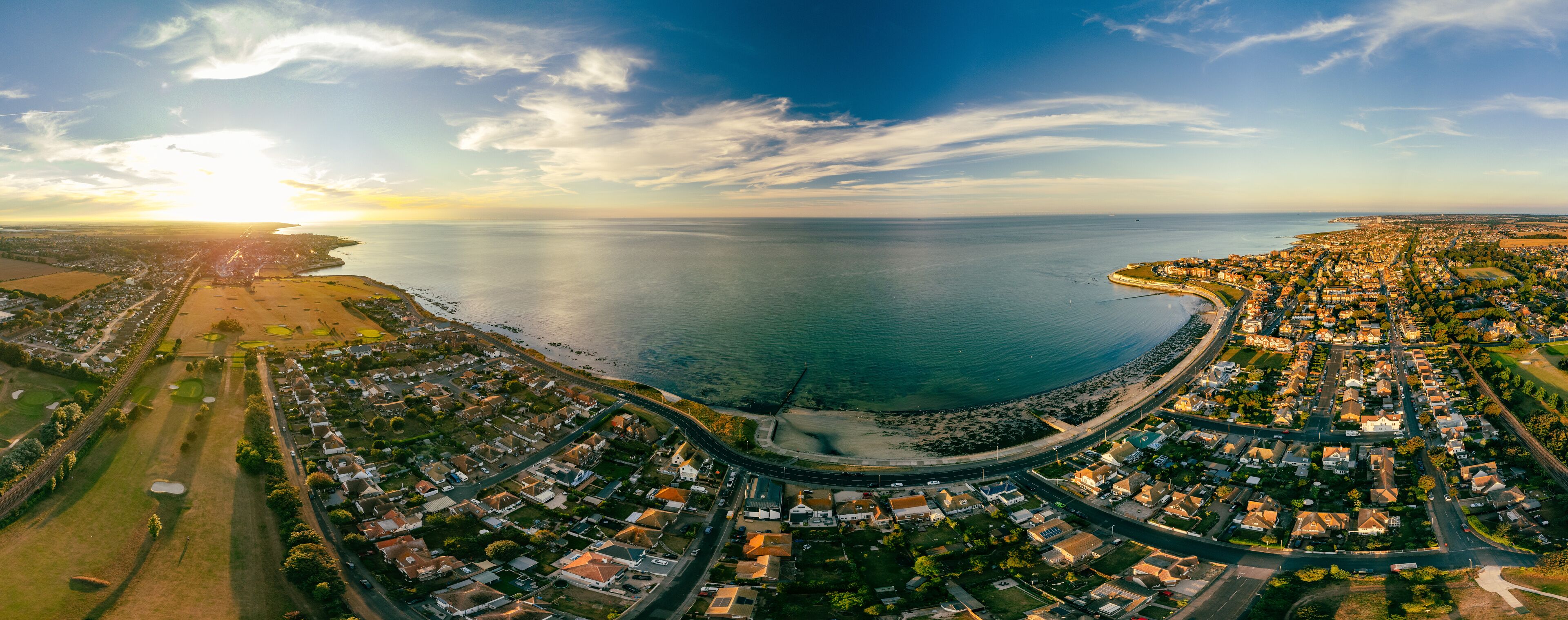 An aerial view of a coastal town with a curved road leading to a beach, surrounded by residential areas and fields.