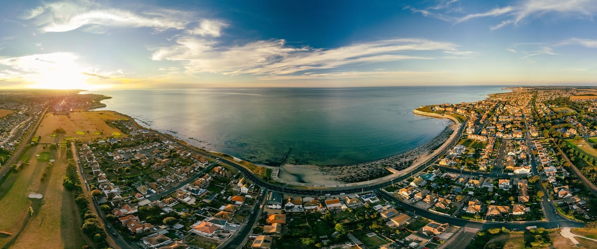 An aerial view of a coastal town with a curved road leading to a beach, surrounded by residential areas and fields.
