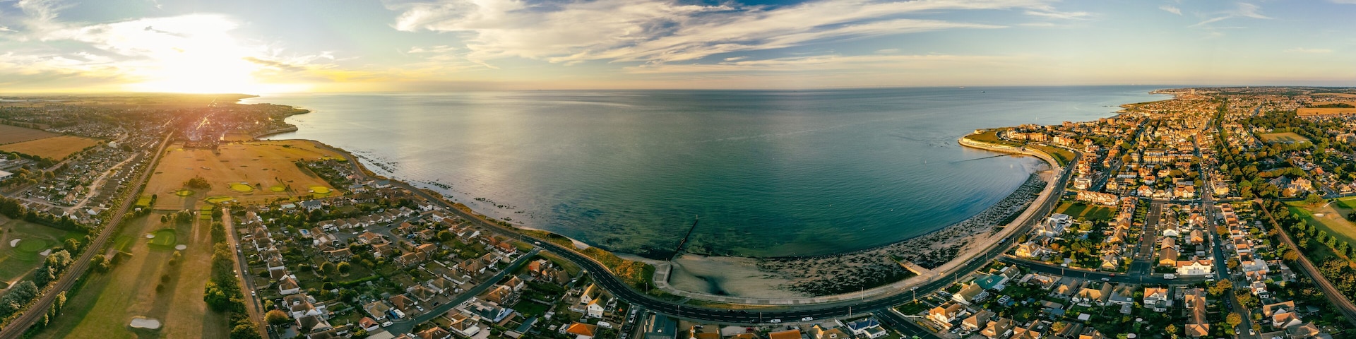 An aerial view of a coastal town with a curved road leading to a beach, surrounded by residential areas and fields.