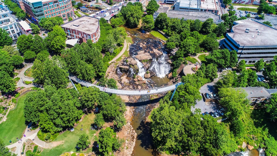 A suspension bridge over a river with waterfalls, surrounded by greenery and urban buildings.