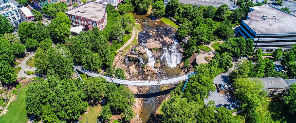 A suspension bridge over a river with waterfalls, surrounded by greenery and urban buildings.