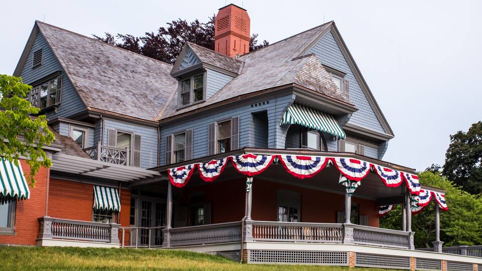 A large, two-story house with a wrap-around porch, red brick walls, and a steeply pitched roof.