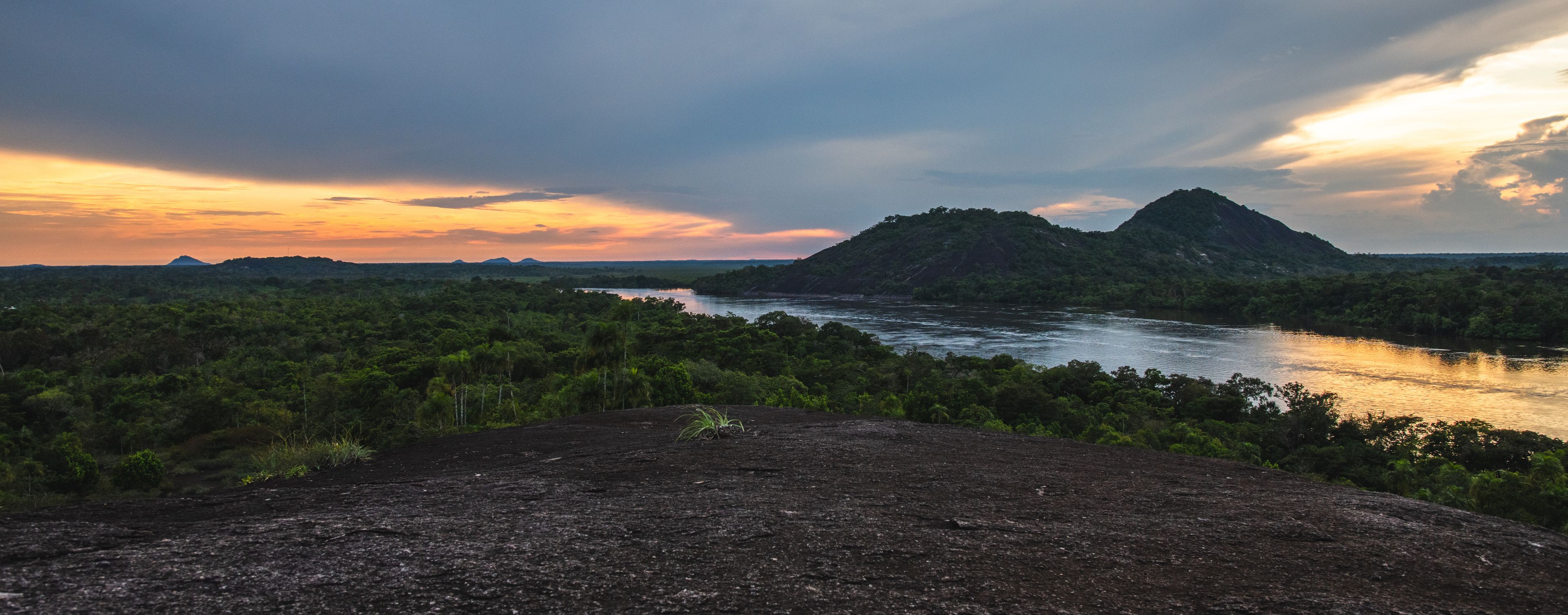 A river winding through a dense forest with a mountainous backdrop during sunset.