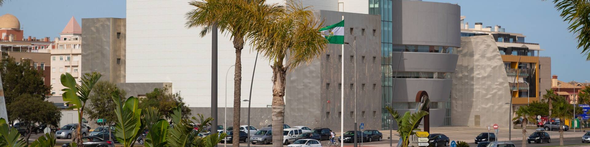 A modern building with a white facade and a glass section, surrounded by palm trees and red flowers.
