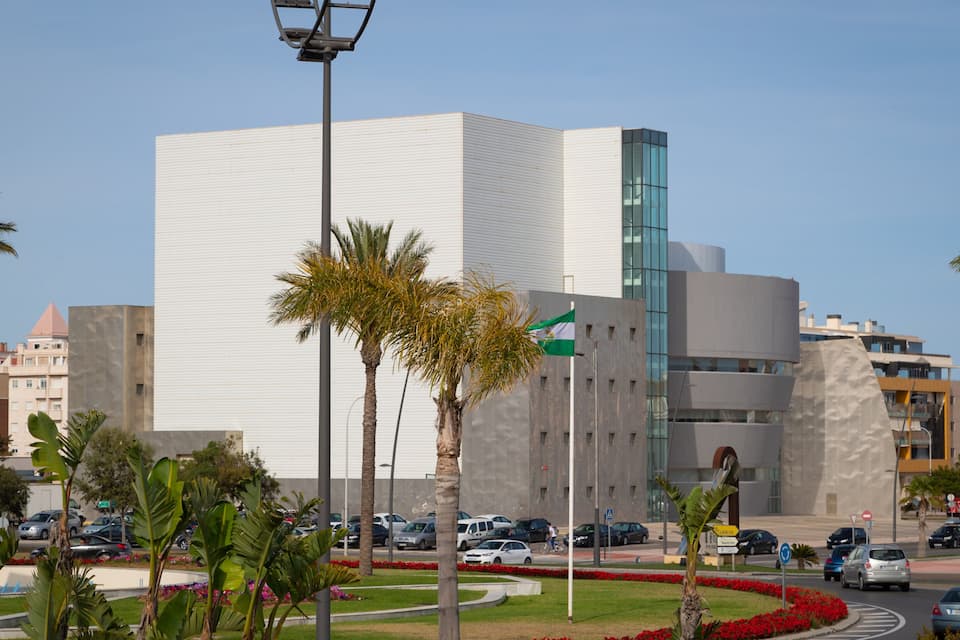 A modern building with a white facade and a glass section, surrounded by palm trees and red flowers.