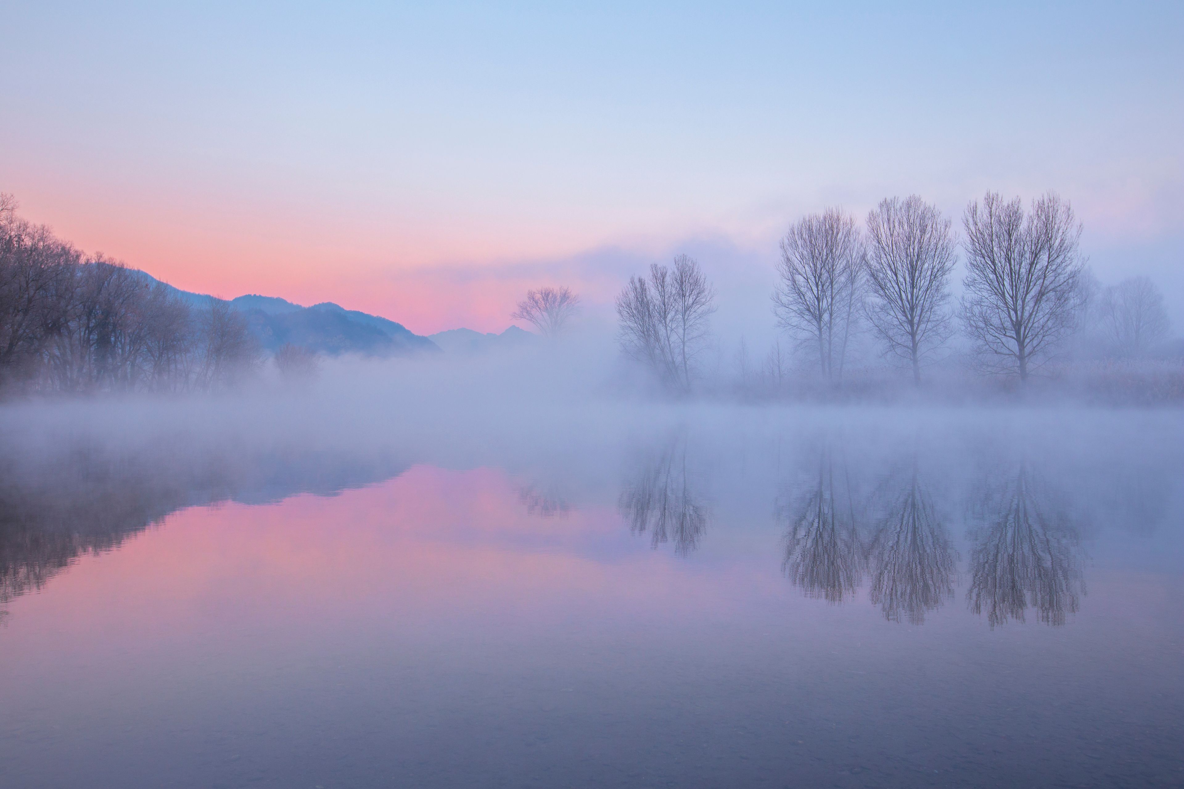 A serene lake with reflections of trees and mountains, surrounded by a misty atmosphere.