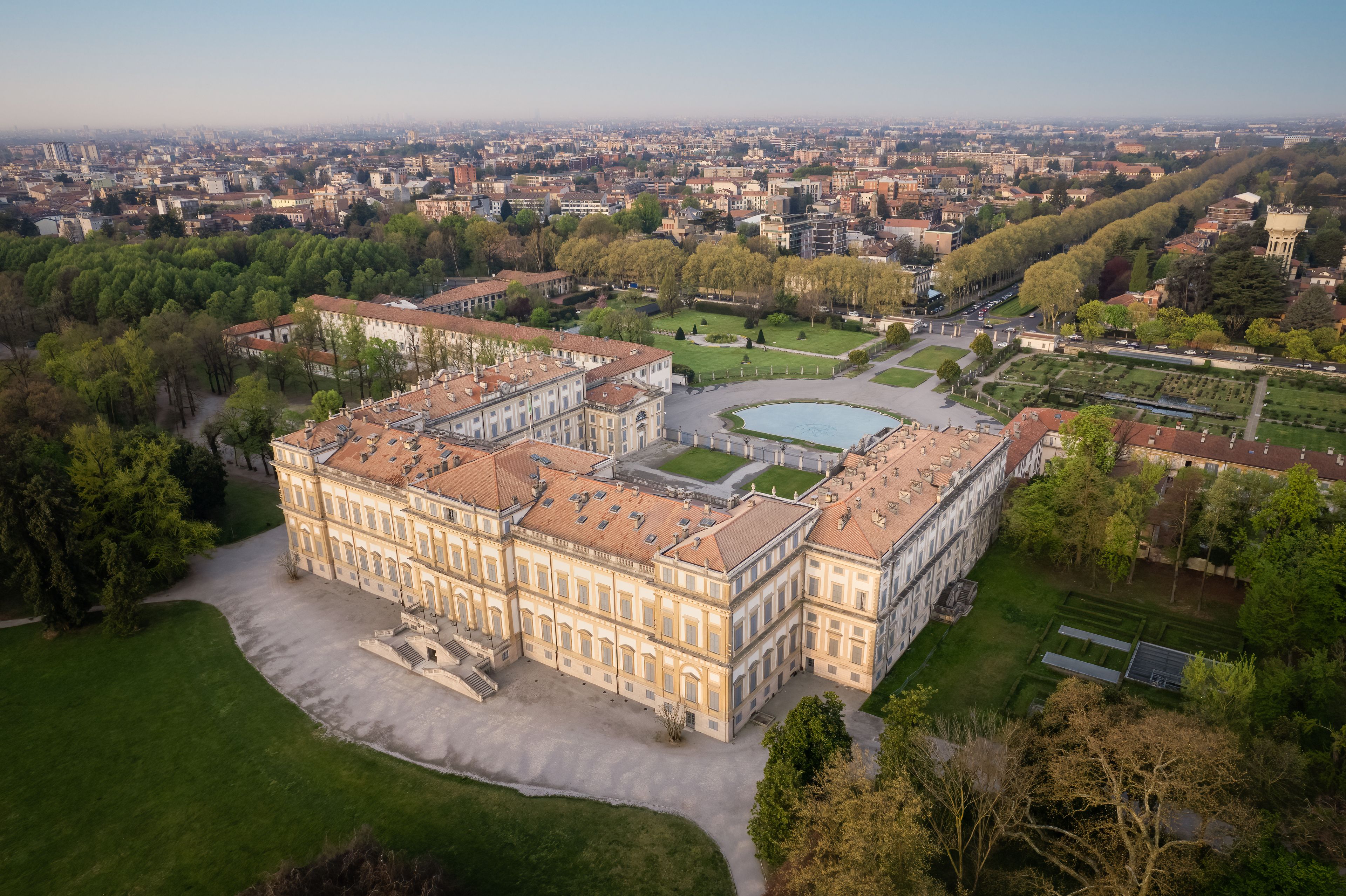 A large historic building with a symmetrical facade, surrounded by well-maintained gardens and a cityscape in the background.