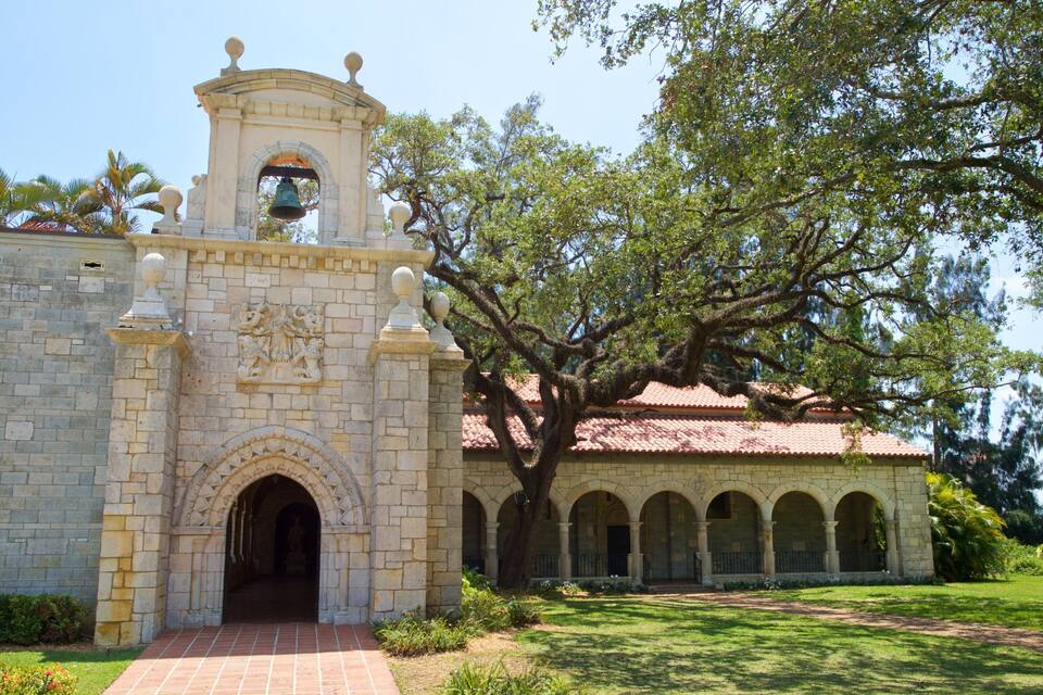 A historic building with a bell tower, arched entrance, and a courtyard with a large tree.