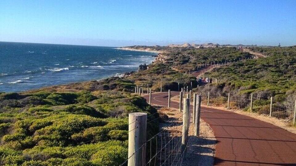 Une route côtière avec une piste rouge, bordée d’une clôture et de végétation, qui mène à une plage.