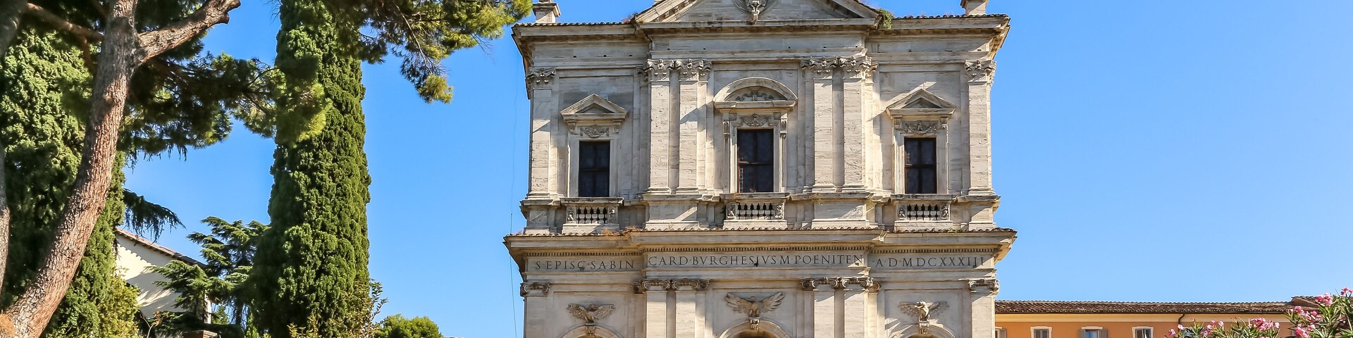 A historic church with a bell tower, surrounded by lush greenery and a clear blue sky.