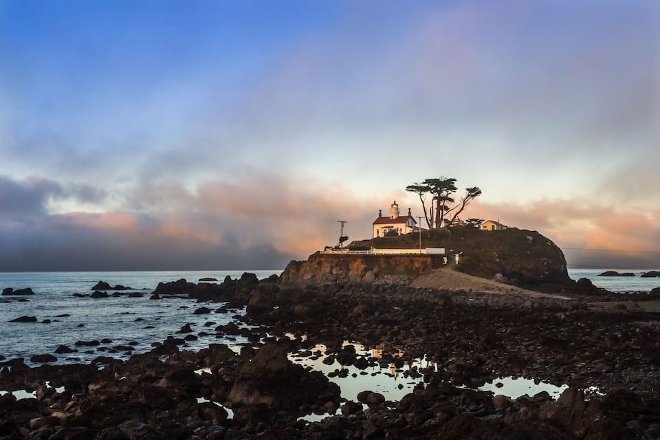 A coastal landscape with rocky shores, a lighthouse, and a white building on a cliff.