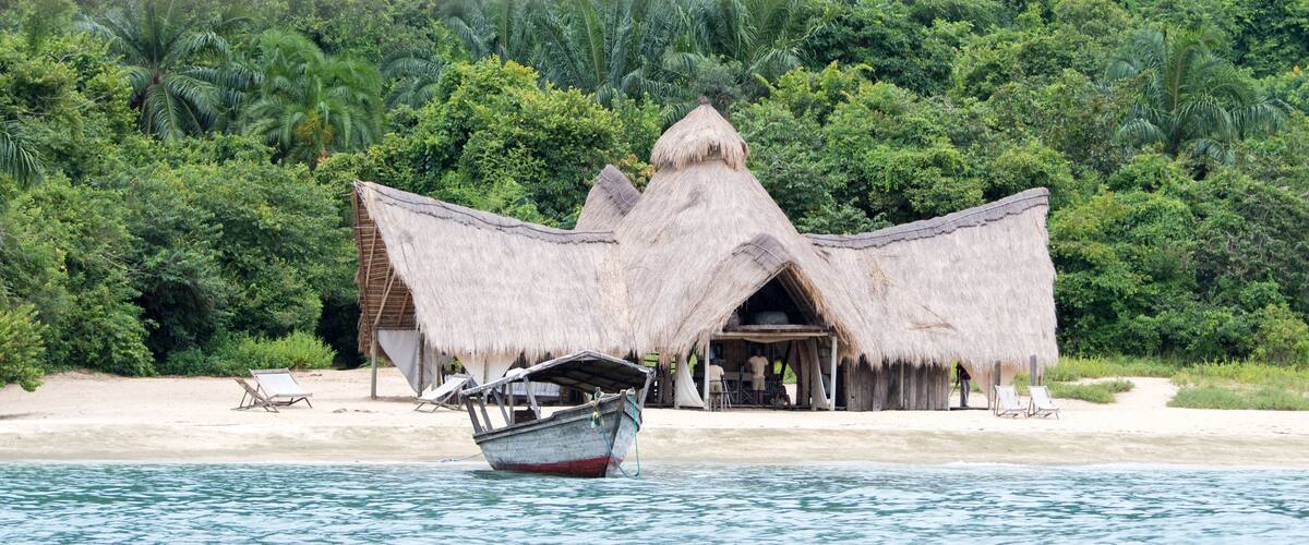 A thatched-roof hut with a boat in the foreground, surrounded by lush greenery and a sandy beach.