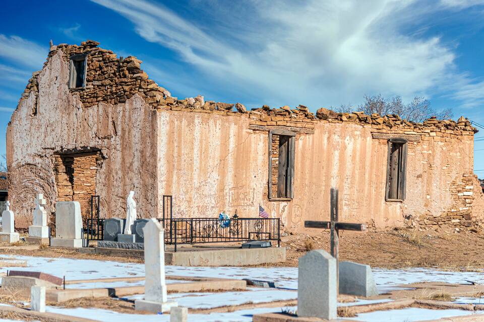 Un vieux bâtiment décrépit, avec un toit en pierre et une petite fenêtre, entouré d’un cimetière où se dressent des pierres tombales et où se trouve une clôture en métal.
