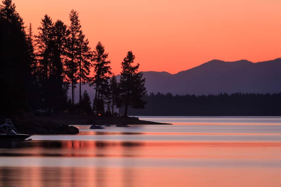A calm lake at sunset with silhouetted trees and mountains in the background.