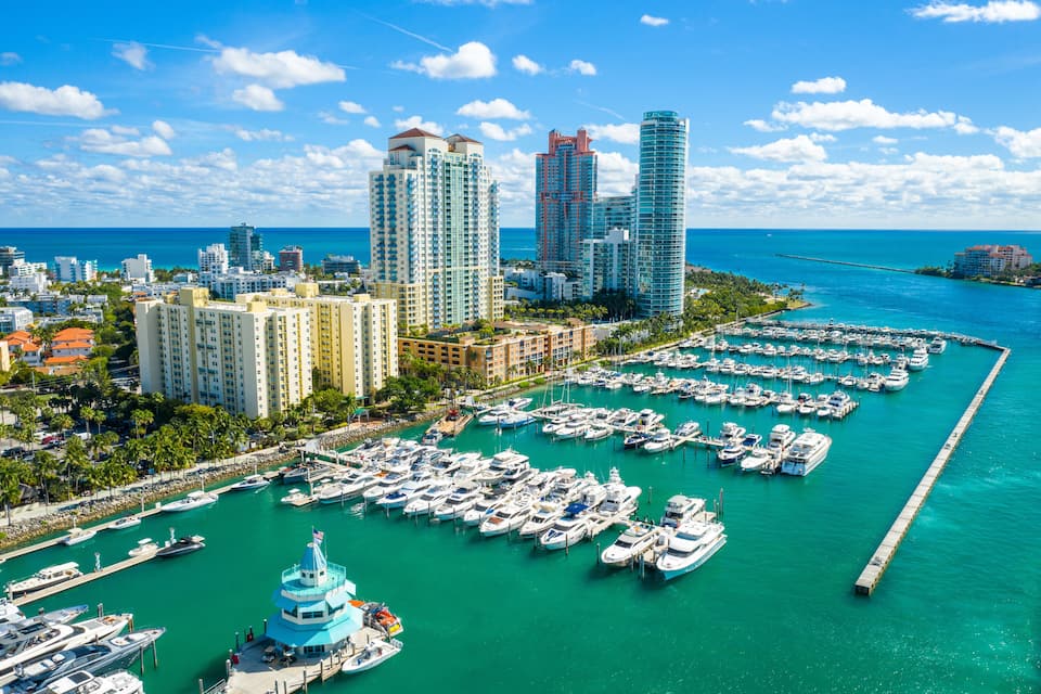 Uma marina com vários barcos atracados, rodeada por edifícios altos e um céu azul límpido.