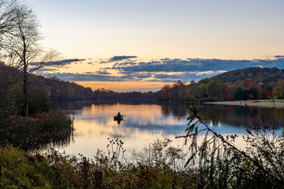 Un lago calmo al tramonto con una barca, circondato da alberi dai colori autunnali e un pendio boscoso.