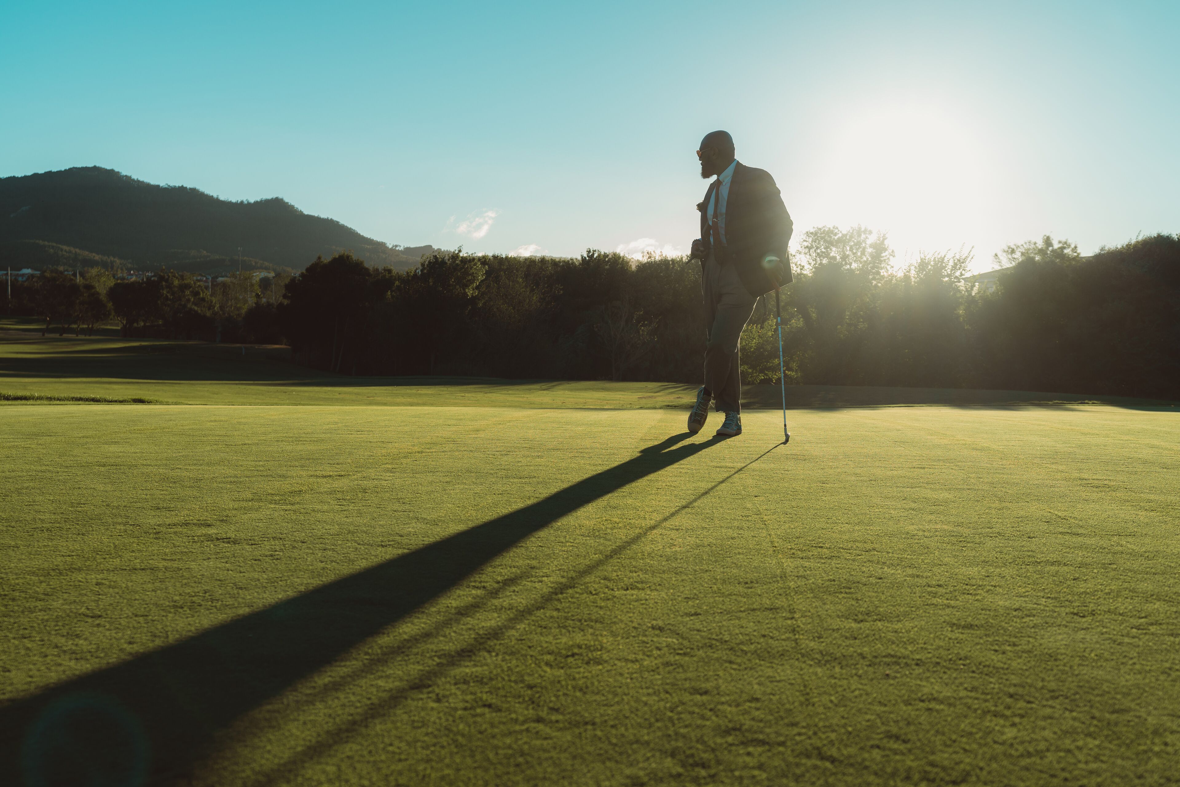 A silhouette of an elegant bald bearded black man in a tailored suit leaning on his gold club while standing on an empty lawn of a golf field, backlit by evening sun casting a long shadow on the grass
